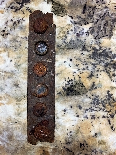 a row of rusted bottle caps on a long flat piece of rusty metal on top of ecoprinted fabric